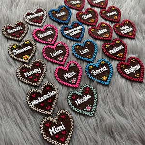 Könnte beinhalten: Eine Sammlung brauner herzförmiger Ornamente mit weiß gestickten Namen wie Tine, Bambi, Luisa, Honey und Mimi. Jedes Herz hat einen dekorativen, gewellten Rand in verschiedenen Farben sowie kleine florale oder herzförmige Akzente.