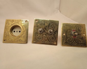 May include: Three decorative brass wall plates with ornate designs. One plate has a white porcelain outlet cover, while the other two have brass toggle switches with red "ON" labels.