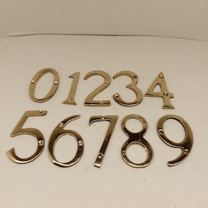 May include: Set of ten gold-toned metal house numbers, 0 through 9. The numbers are individually mounted on a white surface.