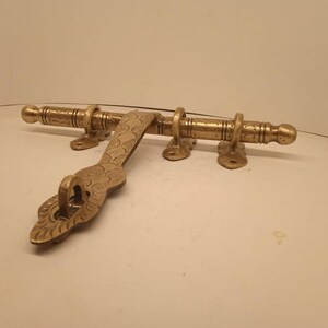 Könnte beinhalten: Ein Vintage-Türriegel aus Messing mit aufwendigen eingravierten Mustern. Der Riegel verfügt über eine horizontale Stange mit drei Ringen und einem dekorativen Griff. Das Messing hat einen warmen, gealterten Ton, der auf einen klassischen oder antiken Stil hindeutet. Diese Hardware ist für Türen geeignet.