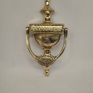 May include: A gold door knocker with an ornate design. The knocker is shaped like a vase with a ring handle. The top of the knocker has a finial.