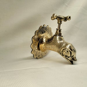 May include: A gold-toned brass faucet shaped like a bird's head with intricate details. The faucet has a decorative backplate with a floral design.
