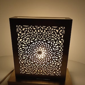 Peut inclure: Une lampe de table de couleur or avec une base carrée et un motif décoratif découpé complexe. La lampe est allumée, projetant une lueur chaude à travers le motif.