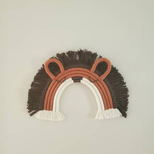 May include: A macrame rainbow wall hanging in the shape of a lion's face. The design features a brown mane, orange ears, and a white and orange striped rainbow. The wall decor is a handmade item.