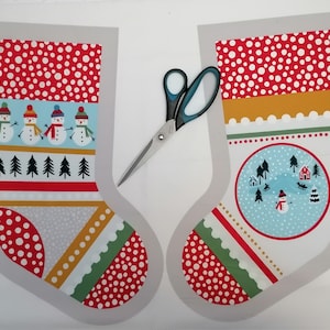 May include: Two Christmas stocking fabric panels with a winter theme. One stocking features snowmen and pine trees, the other a snowy village scene. Red and white polka dot patterns and decorative borders are also present. A pair of scissors is placed between the panels.