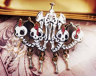 Puede incluir: Pin de esmalte dorado con forma de candelabro esquelético con cuatro portavelas en forma de calavera y colgantes de hueso. El candelabro está decorado con detalles en esmalte rojo.