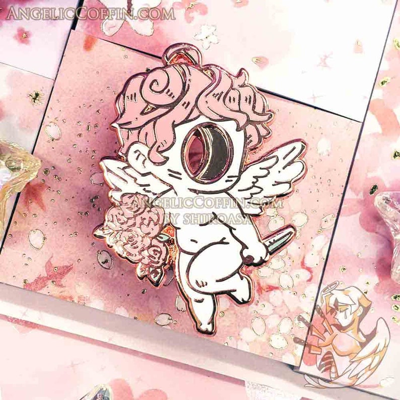 May include: A pink and white enamel pin featuring a cherub with wings, holding a knife and a bouquet of pink roses. The cherub has a single large eye and pink hair. The pin is set against a pink and white background with gold glitter.