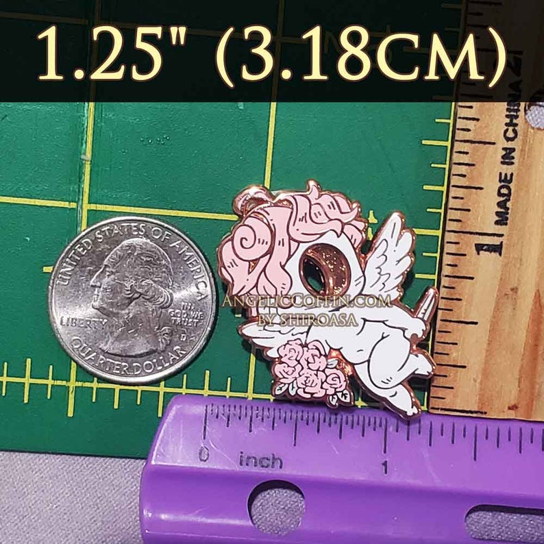 May include: A rose gold enamel pin featuring a cartoon cherub with pink hair, white wings, and a rose bouquet. The pin is 1.25 inches (3.18 cm) in diameter and has the text "ANGELICCOFFIN.COM BY SHIROASA" on the bottom.