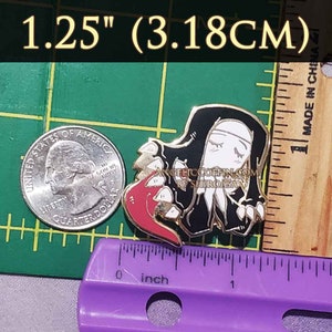 May include: Gold-colored enamel pin featuring a black-haired nun with a red heart in her hand. The pin is 1.25 inches (3.18 cm) in diameter. The text "ANGELICCOFFIN.COM BY SHIROASA" is printed on the pin.