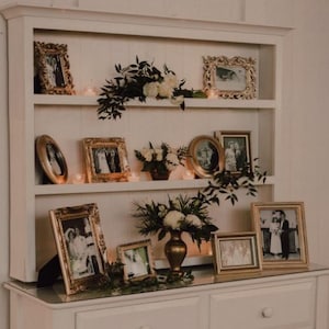 May include: A white cabinet with shelves and drawers, displaying framed photos and floral arrangements. The gold-framed pictures are of various sizes and shapes. The cabinet is decorated with candles and greenery, creating a vintage aesthetic.