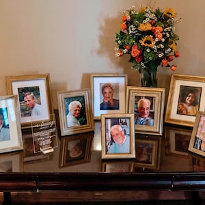 Könnte beinhalten: Eine Sammlung von gerahmten Fotos mit goldfarbenen Rahmen, angeordnet auf einer dunklen Holzoberfläche. Eine transparente Acrylplatte mit Text befindet sich vor den Rahmen. Im Hintergrund steht eine Vase mit Blumen.