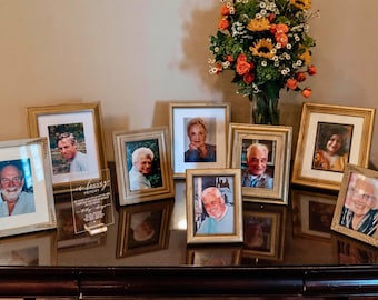 May include: A collection of framed photographs with gold-colored frames, arranged on a dark wooden surface. A clear acrylic plaque with text sits in front of the frames. A vase of flowers is in the background.