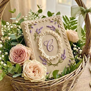 May include: A woven basket overflowing with pink roses, baby's breath, and greenery. A beige book with lavender embroidery and a central "C" monogram sits in the basket. The book's cover features a lace border and lavender sprigs.