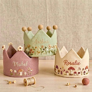 Peut inclure: Trois couronnes en tissu faites à la main dans des tons pastel. L'une est rose poudré avec le nom "Mabel" brodé, une autre est vert sauge avec "Arthur", et la dernière est beige avec "Rosalie". Chaque couronne présente des broderies florales et de champignons.