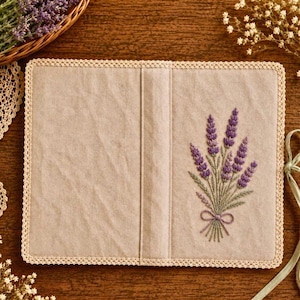 May include: A beige book cover with a lavender embroidery design. The cover features a bouquet of purple lavender tied with a bow. The edges are trimmed with a cream-colored lace.