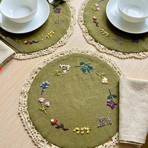 May include: Olive green embroidered placemats with a cream-colored crocheted lace edge. Each placemat features a floral embroidery design in various colors. White bowls and napkins are also visible.