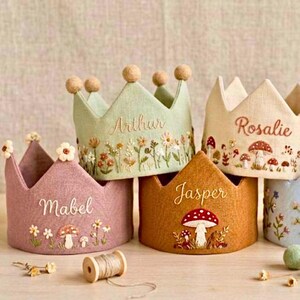 Peut inclure: Plusieurs couronnes en tissu faites à la main, de couleurs rose, vert et beige. Chaque couronne est brodée d'un nom et ornée de motifs floraux et de champignons. Certaines couronnes ont de petites décorations sphériques au sommet.