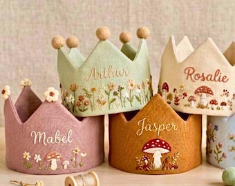 May include: A collection of handmade fabric crowns in various colors, including pink, green, and tan. Each crown is embroidered with a name and decorative elements like flowers and mushrooms. Some crowns have small, round embellishments at the points.