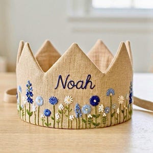 Peut inclure: Une couronne en tissu beige brodée du nom "Noah" en bleu marine. La couronne est ornée de broderies florales bleues, blanches et vertes. La couronne a une sangle de chaque côté.