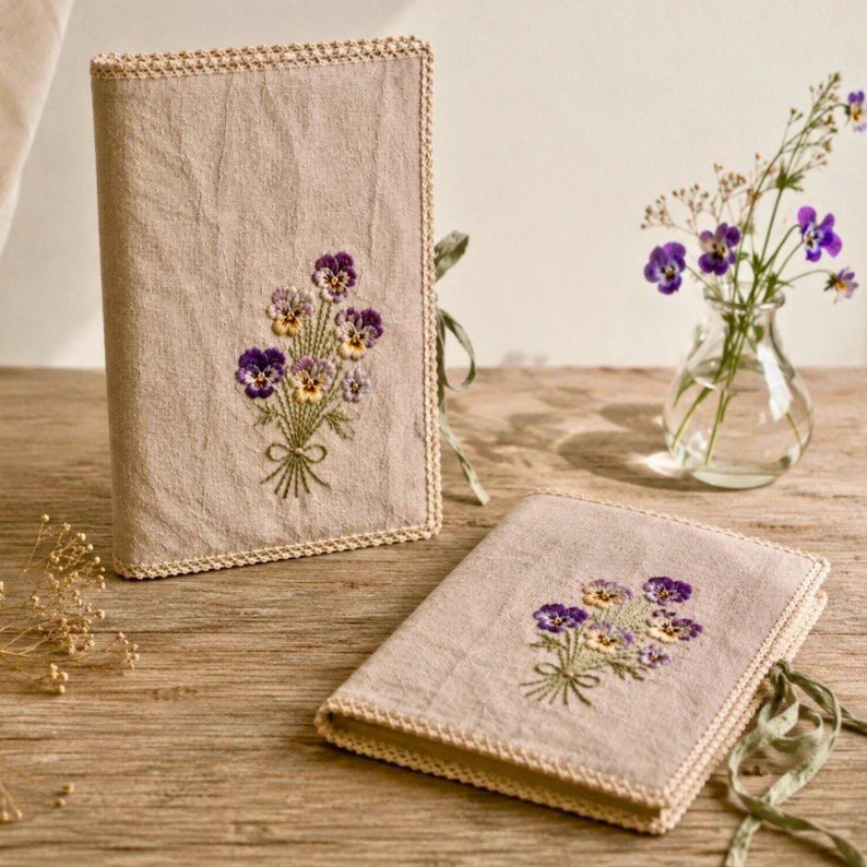 May include: Two beige book covers with embroidered purple and yellow pansies. The covers have a decorative border and green ribbon ties. One cover is upright, and the other is lying flat on a wooden surface.