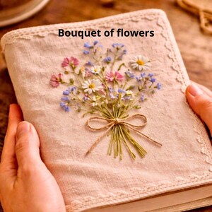May include: A beige linen journal with a floral embroidery of pink, blue, and white flowers tied with twine. The text "Bouquet of flowers" is above the embroidery. The journal has a lace trim.