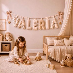 Peut inclure: Une chambre d'enfant décorée dans des tons beiges doux, avec une bannière "VALERIA" au-dessus d'un lit en bois. On y trouve un lapin en peluche, une voiture en bois et un service à thé. Une petite fille joue avec le service à thé sur un tapis moelleux.