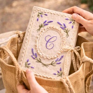 May include: A beige journal with lavender embroidery and a large embroidered letter "C" in a decorative oval. The journal is trimmed with lace and tied with a ribbon. The journal is held in a burlap bag.