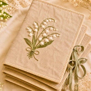 May include: A stack of beige linen napkins with embroidered lily of the valley floral design. The napkins have a decorative border and are tied with a green ribbon. The embroidery features white flowers and green leaves.