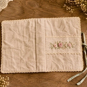 May include: Beige linen journal cover with a floral embroidery design. The cover features a scalloped lace trim and a pocket with embroidered pink roses and small white flowers. A green ribbon is attached for closure.
