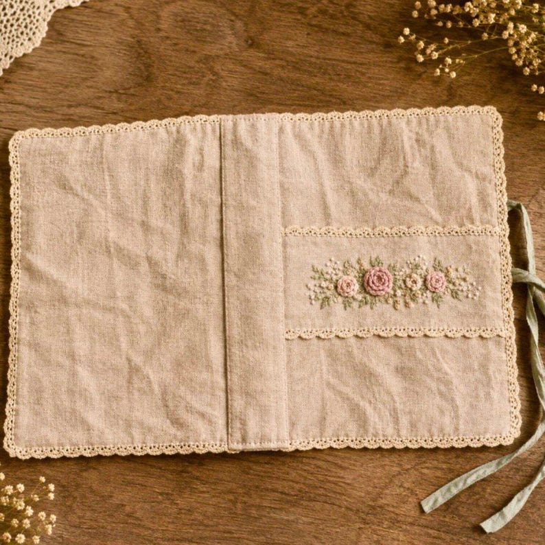May include: Beige linen journal cover with a floral embroidery design. The cover features a scalloped lace trim and a pocket with embroidered pink roses and small white flowers. A green ribbon is attached for closure.