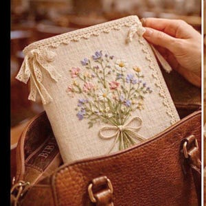 May include: A beige book cover with an embroidered floral design in pastel colors. The cover is trimmed with lace and tied with delicate ribbons. The book is inside a brown leather bag.