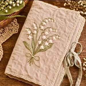 May include: A beige linen journal with a detailed embroidered lily of the valley design. The cover features a bouquet of white flowers with green leaves and a delicate bow. The edges are trimmed with cream-colored lace, and a sage green ribbon ties the journal closed.