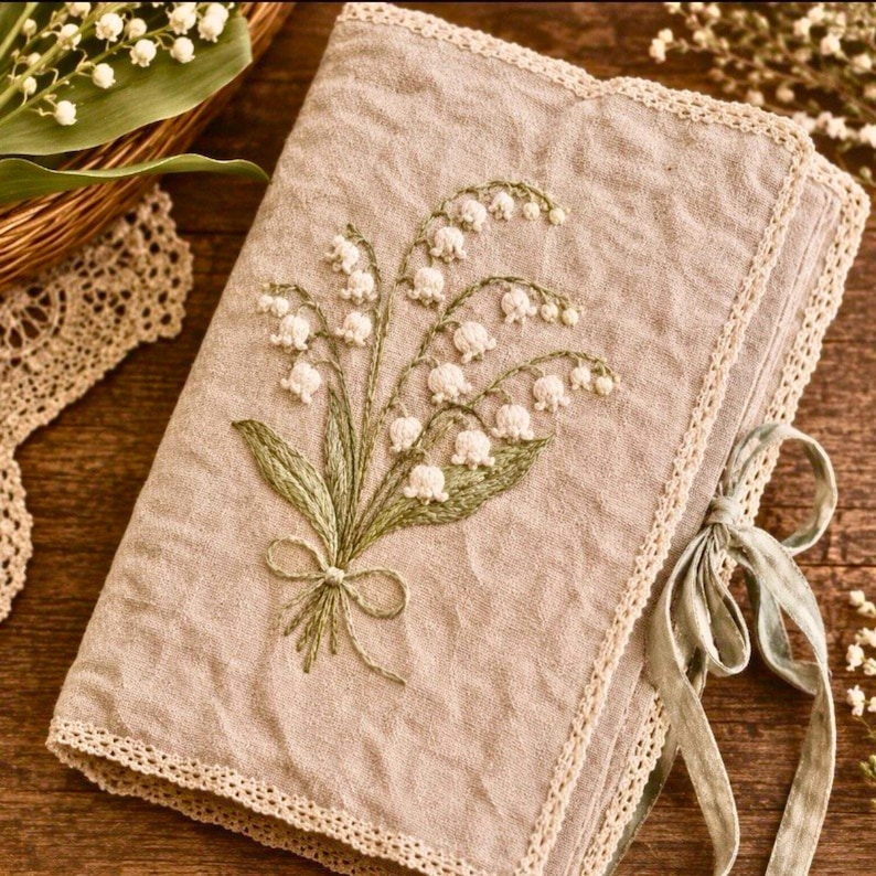 May include: A beige linen journal with a detailed embroidered lily of the valley design. The cover features a bouquet of white flowers with green leaves and a delicate bow. The edges are trimmed with cream-colored lace, and a sage green ribbon ties the journal closed.