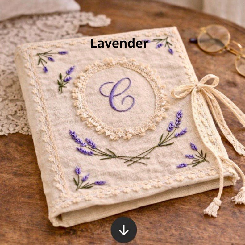 May include: A beige linen journal with embroidered lavender sprigs and a central embroidered "C". The journal is trimmed with lace and tied with a ribbon. The word "Lavender" is printed on the top.