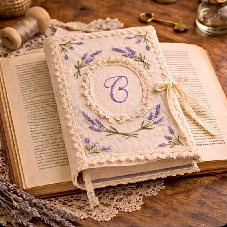 May include: A cream-colored journal with a lavender embroidered design and the letter "C" in the center. The journal is open, revealing pages, and is adorned with lace trim and a ribbon tie. The cover features sprigs of lavender and a decorative border.