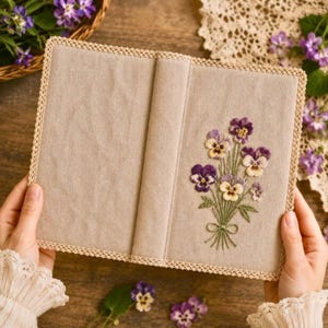 May include: Beige book cover with a floral embroidery of purple and yellow pansies. The cover has a decorative lace trim and a stitched spine. The embroidery features green stems and leaves, tied with a green bow.
