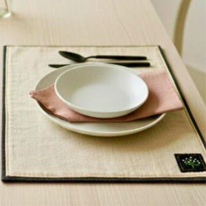 May include: A dining table setting with a white bowl and plate on a pink napkin. The setting is on a beige placemat with a dark border, with black cutlery. A small embroidered detail is on the placemat.