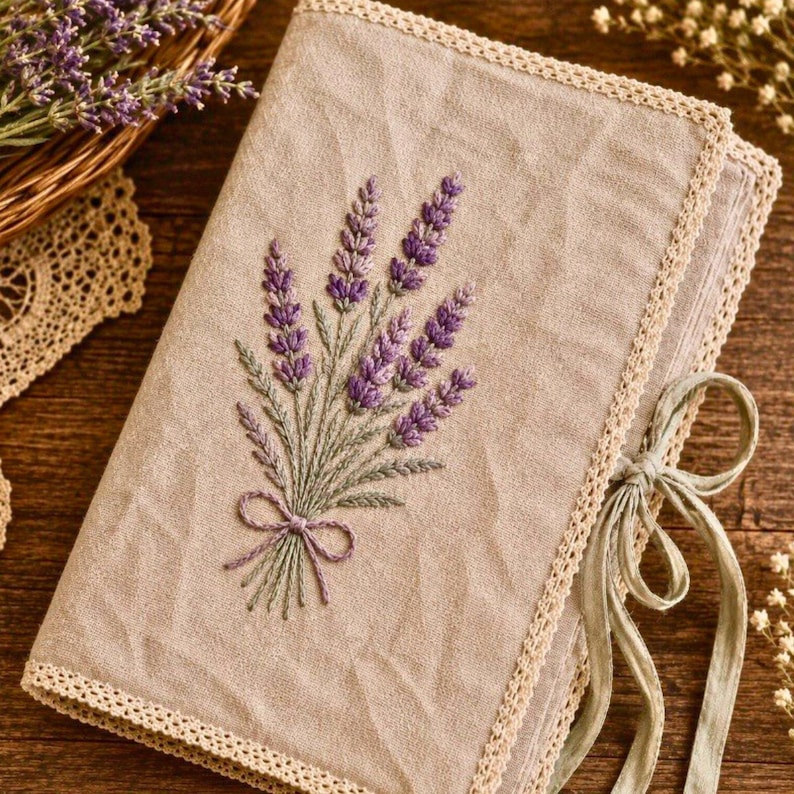 May include: A beige fabric journal cover with a lavender embroidery design. The embroidered lavender bouquet is purple and green, tied with a small bow. The cover is trimmed with a cream-colored lace and tied with a green ribbon.