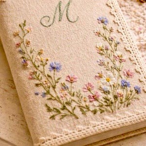 May include: A beige journal with embroidered floral designs in pink, blue, and yellow. The letter "M" is embroidered in green at the top. The journal has a lace trim along the edge.