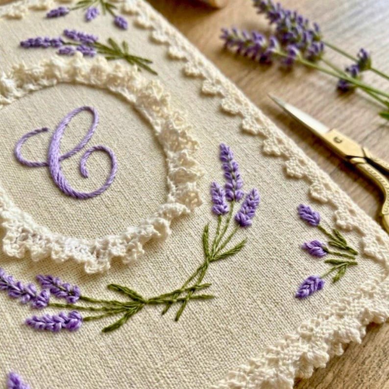 May include: Cream-colored fabric with embroidered lavender sprigs and a letter "C" in purple thread. The edges are trimmed with a delicate lace border. A pair of gold scissors rests nearby, suggesting a craft or sewing project.