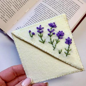 May include: A handmade, off-white felt bookmark with embroidered purple lavender flowers and green stems. The bookmark is triangular and has a stitched border. A book is open in the background.