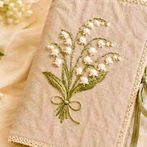 May include: A beige linen journal cover with an embroidered bouquet of white lily of the valley flowers and green leaves. The embroidery is detailed, with a green bow at the base of the bouquet. The cover has a lace trim and a green ribbon tie.