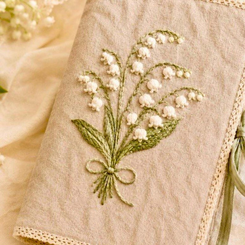 May include: A beige linen journal cover with an embroidered bouquet of white lily of the valley flowers and green leaves. The embroidery is detailed, with a green bow at the base of the bouquet. The cover has a lace trim and a green ribbon tie.