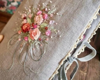 May include: A gray linen journal with a floral embroidery design. The embroidery features pink, red, and peach roses with green leaves and delicate white accents. The journal is trimmed with cream lace and tied with a gray velvet ribbon.