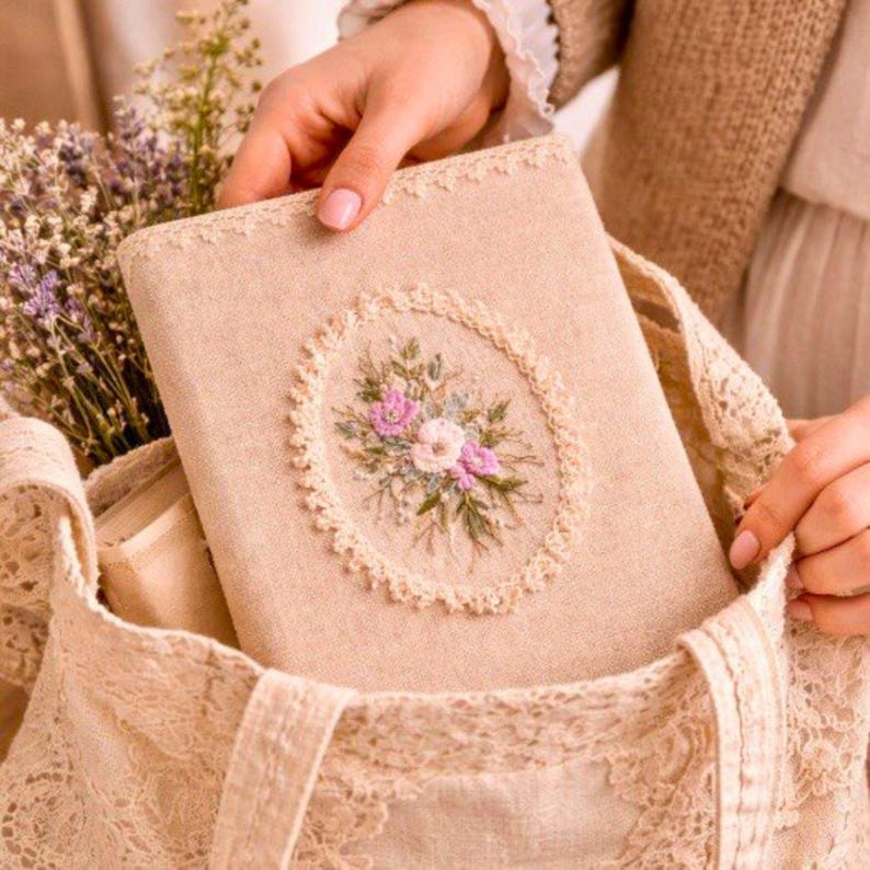 May include: A beige journal with an embroidered floral design in a lace-trimmed tote bag. The journal cover features a central oval of pink and white flowers with green leaves. The tote bag is made of lace.