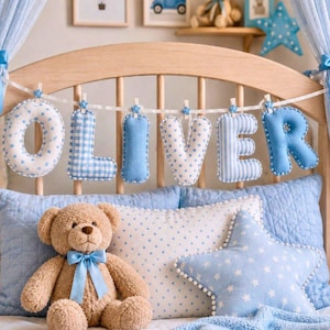 May include: A nursery scene with a light brown teddy bear wearing a blue bow, blue pillows, and a star-shaped pillow. The name "OLIVER" is spelled out in blue fabric letters, each with a different pattern, hanging above the bed.