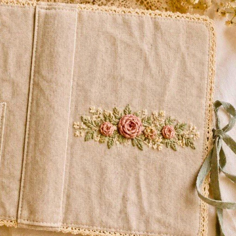 May include: Beige fabric journal cover with embroidered pink roses and green leaves. The cover has a lace trim and a sage green ribbon tie. The embroidery is centered on the front of the cover.
