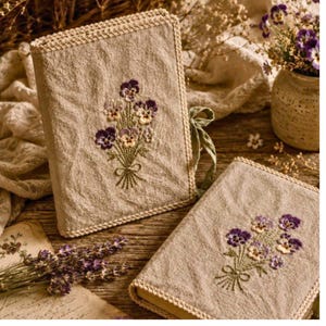 May include: Two beige linen book covers with embroidered purple and yellow pansies. The covers have a crocheted border and a green ribbon tie. One book is upright, the other is lying flat.