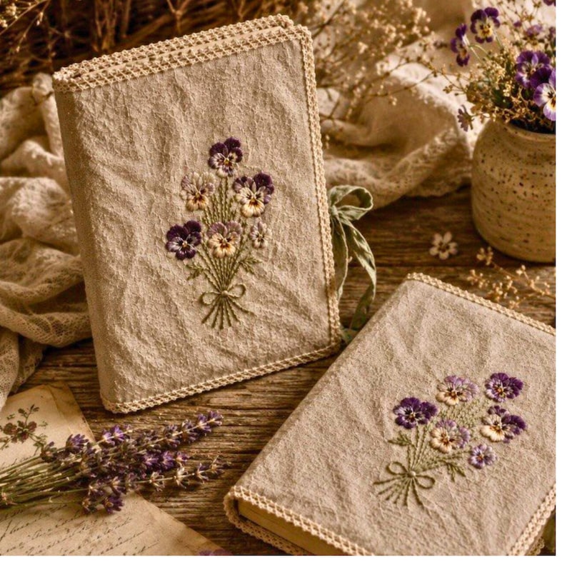 May include: Two beige linen book covers with embroidered purple and yellow pansies. The covers have a crocheted border and a green ribbon tie. One book is upright, the other is lying flat.