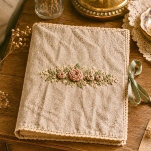 May include: A beige linen journal with embroidered pink roses and greenery. The cover is trimmed with lace and a sage green ribbon. The journal is open, revealing the pages within. The overall aesthetic is vintage and handcrafted.